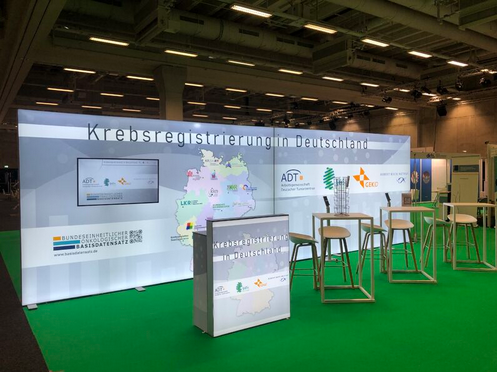 Zu sehen ist der gemeinsame Stand inmitten der Messe. Man sieht einen Standpult und Stühle an Tischen.